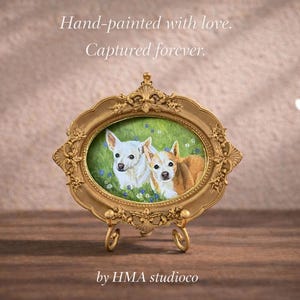 May include: An oval-shaped gold-toned ornate frame containing a painting of two small dogs in a green field with blue flowers. The frame rests on a matching gold-toned metal stand. The text "Hand-painted with love. Captured forever." and "by HMA studioco" appear.