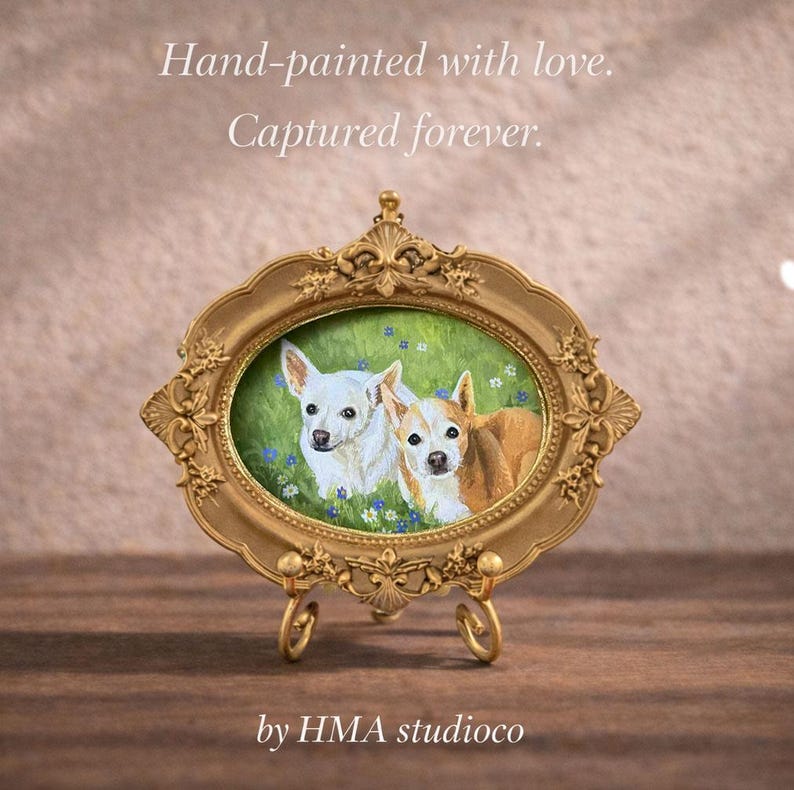 May include: An oval-shaped gold-toned ornate frame containing a painting of two small dogs in a green field with blue flowers. The frame rests on a matching gold-toned metal stand. The text "Hand-painted with love. Captured forever." and "by HMA studioco" appear.