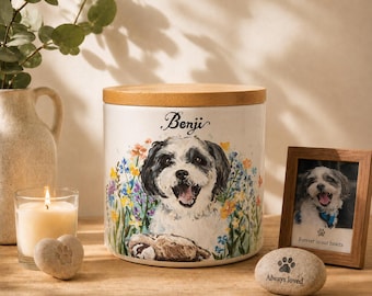 May include: A cylindrical white ceramic pet memorial urn with a wooden lid, featuring a painted illustration of a dog named Benji surrounded by colorful wildflowers. The urn is displayed alongside a small framed photo and a stone with a paw print.