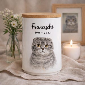 May include: A white ceramic cylindrical jar with a wooden lid, featuring a detailed illustration of a grey tabby cat with yellow eyes. The text "Franceschi" is printed in black script above the dates "2011 - 2022" in a simple black font.