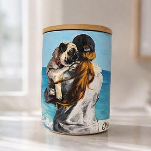 May include: A cylindrical ceramic canister with a bamboo lid featuring a painted scene of a person with long reddish-brown hair wearing a black cap, holding a fawn-colored pug against a blue ocean backdrop. The name Otis is written in script.