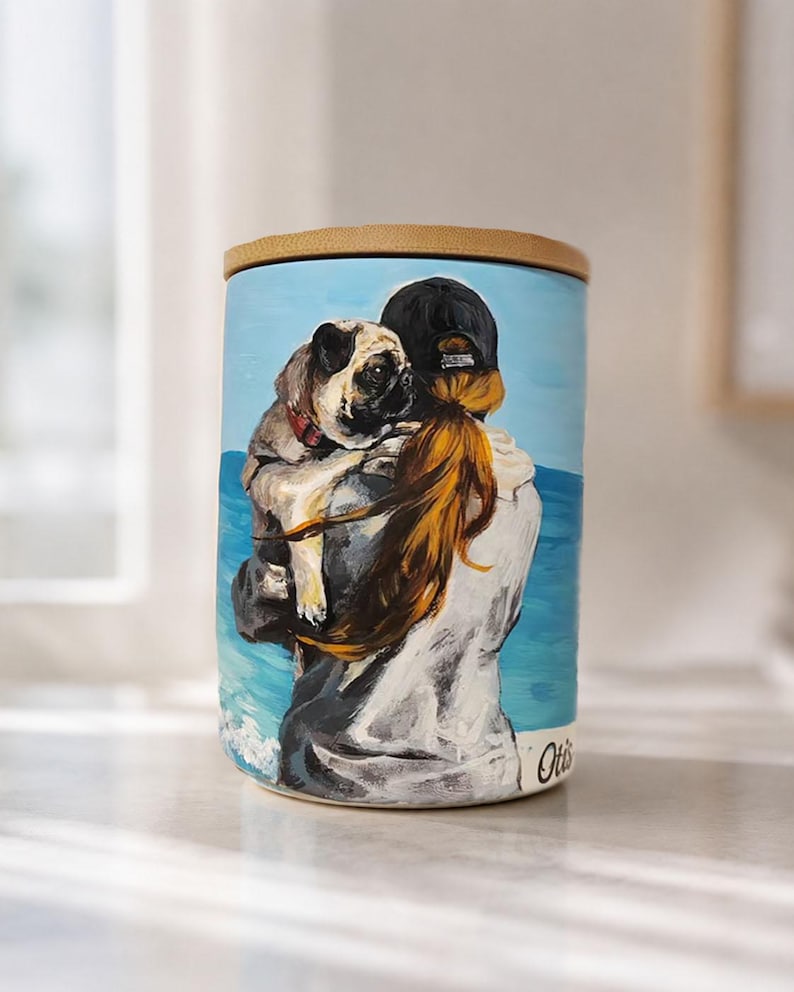 May include: A cylindrical ceramic canister with a bamboo lid featuring a painted scene of a person with long reddish-brown hair wearing a black cap, holding a fawn-colored pug against a blue ocean backdrop. The name Otis is written in script.