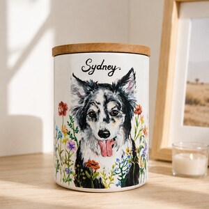 May include: A cylindrical white ceramic canister with a wooden lid featuring a painted illustration of a black and white dog with its tongue out, surrounded by colorful wildflowers, with the name Sydney written in black script above the dog.