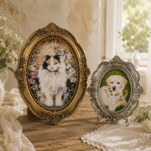 May include: Two oval-shaped picture frames containing pet portraits. The larger frame is gold with ornate floral detailing and holds a painting of a calico cat. The smaller frame is silver with intricate scrollwork and holds a painting of a dog.