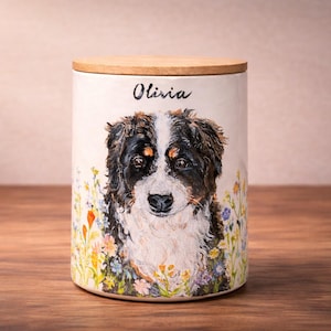 May include: A cylindrical ceramic canister with a light-colored finish, featuring a painted portrait of a Bernese Mountain Dog surrounded by colorful wildflowers. The name Olivia is printed in black script above the dog. It has a wooden lid.