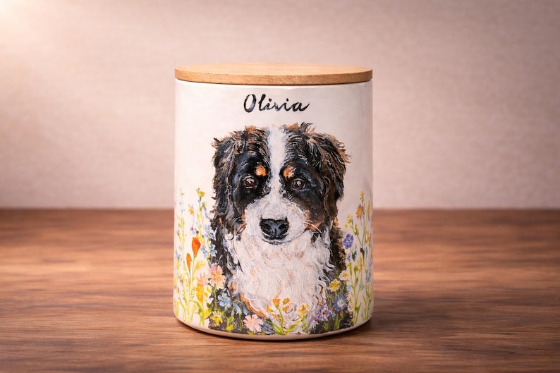 May include: A cylindrical ceramic canister with a light-colored finish, featuring a painted portrait of a Bernese Mountain Dog surrounded by colorful wildflowers. The name Olivia is printed in black script above the dog. It has a wooden lid.