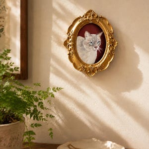 May include: An oval-shaped gold-colored picture frame with ornate, embossed floral detailing along the edges. The frame contains a portrait of a light-colored cat with blue eyes set against a deep red background, mounted on a textured wall.