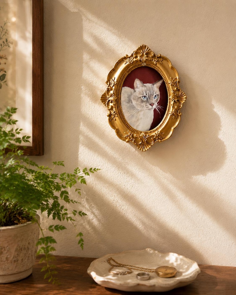 May include: An oval-shaped gold-colored picture frame with ornate, embossed floral detailing along the edges. The frame contains a portrait of a light-colored cat with blue eyes set against a deep red background, mounted on a textured wall.