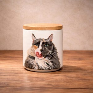 May include: A cylindrical white ceramic canister featuring a detailed painting of a long-haired calico cat licking its nose. The container is topped with a circular, light-colored wooden lid that fits flush with the top rim of the vessel.