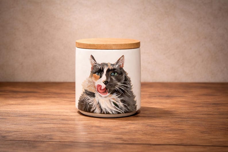 May include: A cylindrical white ceramic canister featuring a detailed painting of a long-haired calico cat licking its nose. The container is topped with a circular, light-colored wooden lid that fits flush with the top rim of the vessel.