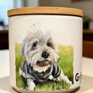 May include: A white ceramic canister with a bamboo lid featuring a hand-painted portrait of a fluffy white dog wearing a dark gray sweater, set against a green grassy background. The name Griffin is printed in black lettering on the side.