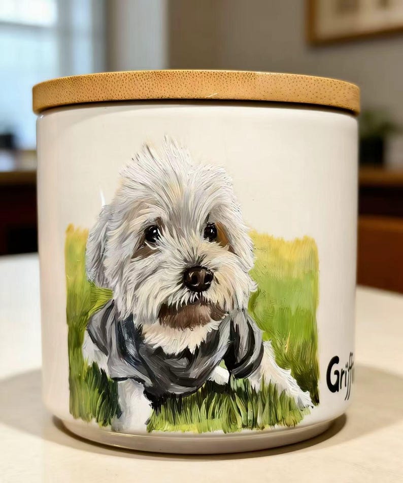May include: A white ceramic canister with a bamboo lid featuring a hand-painted portrait of a fluffy white dog wearing a dark gray sweater, set against a green grassy background. The name Griffin is printed in black lettering on the side.