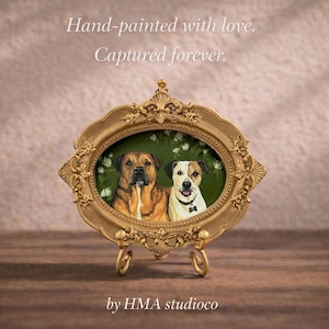 May include: An oval gold-toned ornate frame containing a painting of two dogs, one brown and one white with brown patches, against a green floral background. The text "Hand-painted with love. Captured forever." and "by HMA studioco" is visible.