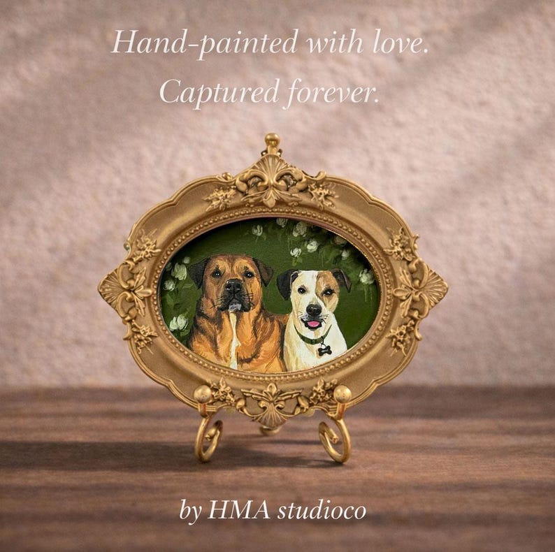 May include: An oval gold-toned ornate frame containing a painting of two dogs, one brown and one white with brown patches, against a green floral background. The text "Hand-painted with love. Captured forever." and "by HMA studioco" is visible.
