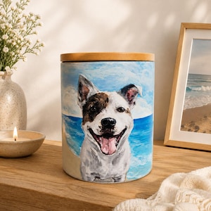 May include: A cylindrical ceramic canister featuring a hand-painted portrait of a smiling dog with white and brown fur. The background depicts a beach scene with blue ocean waves and sandy shores. The container is topped with a round, light wood lid.