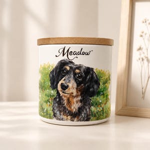 May include: A white ceramic cylindrical canister with a wooden lid, featuring a painted illustration of a black and tan long-haired dachshund in a green meadow. The name Meadow is printed in black cursive script above the dog illustration.