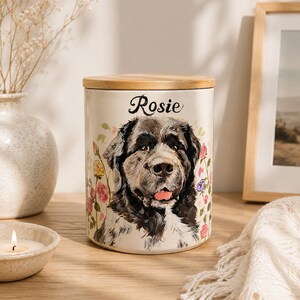 May include: A cylindrical ceramic canister with a light cream finish and a wooden lid. The front features a painted portrait of a black and white dog with the name Rosie printed in black script above the dog, surrounded by colorful floral accents.