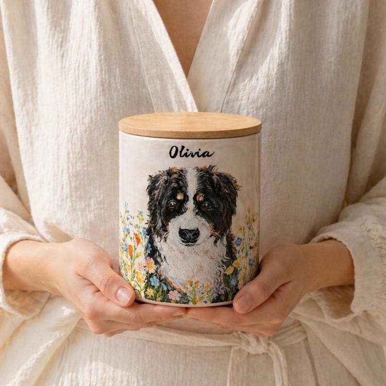 May include: A cylindrical ceramic canister with a light beige finish, featuring a painted portrait of a black, white, and brown dog surrounded by colorful wildflowers. The name Olivia is printed in black script above the dog. It has a wooden lid.