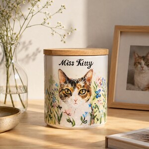 May include: A cylindrical ceramic canister with a light-colored finish, featuring a painted calico cat surrounded by colorful wildflowers. The text "Miss Kitty" is printed in black script above the cat. It has a flat, circular wooden lid.