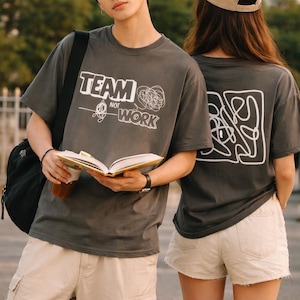 May include: Two people wearing charcoal gray t-shirts with white graphic designs. One shirt says "TEAM NOT WORK" and the other has an abstract design. The person on the left is holding a book and a drink, wearing tan cargo pants.