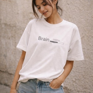 May include: White t-shirt with the words "Brain Loading" printed in black. The shirt is paired with blue jeans. The model is standing in front of a neutral-colored wall.
