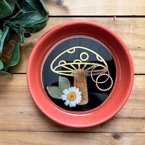 May include: A red ceramic trinket dish with a black interior and a gold mushroom design. The dish has a white daisy and green leaves inside. Two gold rings are placed inside the dish.