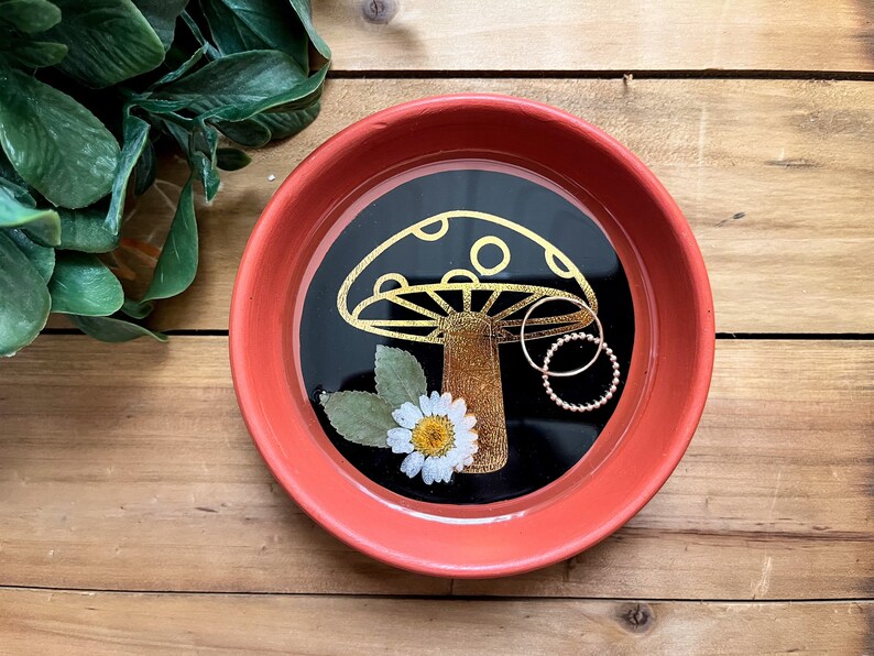 May include: A red ceramic trinket dish with a black interior and a gold mushroom design. The dish has a white daisy and green leaves inside. Two gold rings are placed inside the dish.