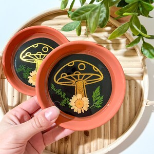 May include: Two terracotta coasters with black resin and gold mushroom designs. Each coaster features a pressed flower and greenery.