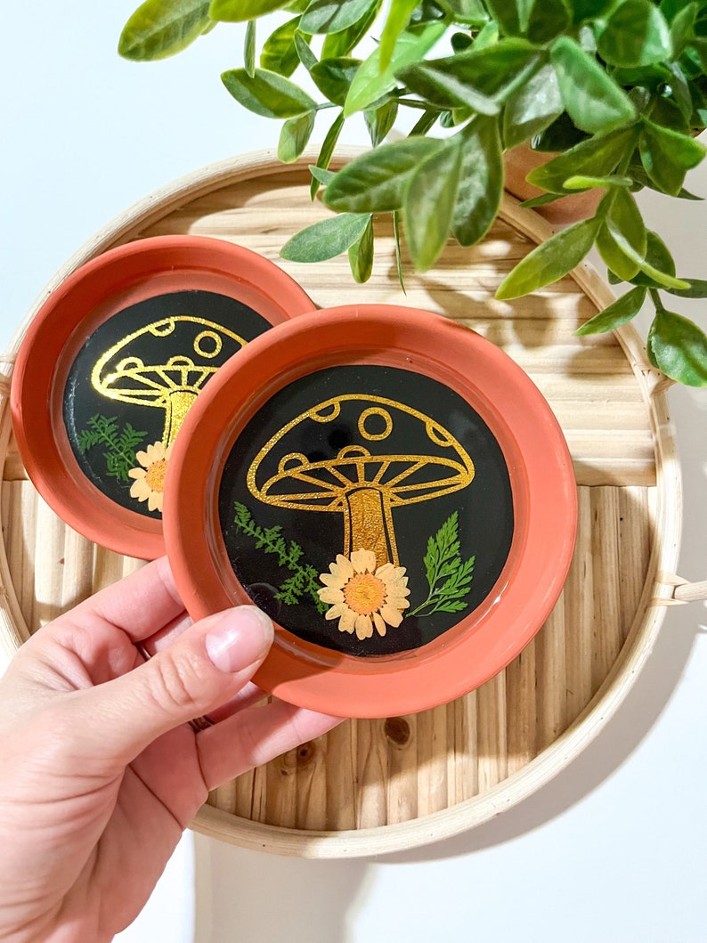 May include: Two terracotta coasters with black resin and gold mushroom designs. Each coaster features a pressed flower and greenery.