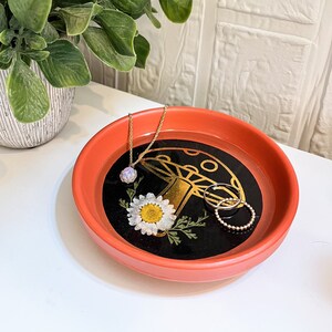 May include: A red ceramic trinket dish with a black and gold mushroom design and a white daisy. The dish is filled with a gold necklace and two gold rings.