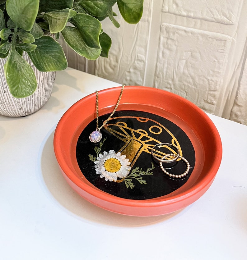 May include: A red ceramic trinket dish with a black and gold mushroom design and a white daisy. The dish is filled with a gold necklace and two gold rings.