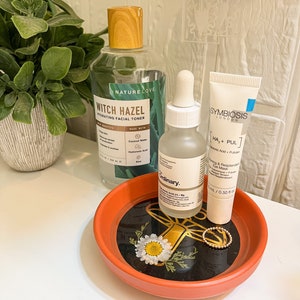 May include: Three skincare products on a small orange tray with a black and gold floral design. The products are a clear bottle of witch hazel toner, a clear glass bottle of hyaluronic acid serum, and a white tube of a hydrating face mask. The toner bottle has a wooden lid and the text "Nature Love Witch Hazel Hydrating Facial Toner" on the label. The serum bottle has a dropper and the text "The Ordinary Hyaluronic Acid 2% + B5" on the label. The face mask tube has the text "Symbiosis London HA + PUL" on the label.