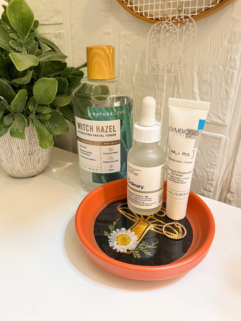 May include: Three skincare products on a small orange tray with a black and gold floral design. The products are a clear bottle of witch hazel toner, a clear glass bottle of hyaluronic acid serum, and a white tube of a hydrating face mask. The toner bottle has a wooden lid and the text "Nature Love Witch Hazel Hydrating Facial Toner" on the label. The serum bottle has a dropper and the text "The Ordinary Hyaluronic Acid 2% + B5" on the label. The face mask tube has the text "Symbiosis London HA + PUL" on the label.