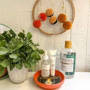 May include: A small orange ceramic dish with a floral design holds three skincare products: a clear glass bottle with a dropper labelled "The Ordinary", a white tube labelled "Symbiosis HA + PL", and a clear glass bottle with a brown cap labelled "Nature's Love Witch Hazel Hydrating Facial Toner".