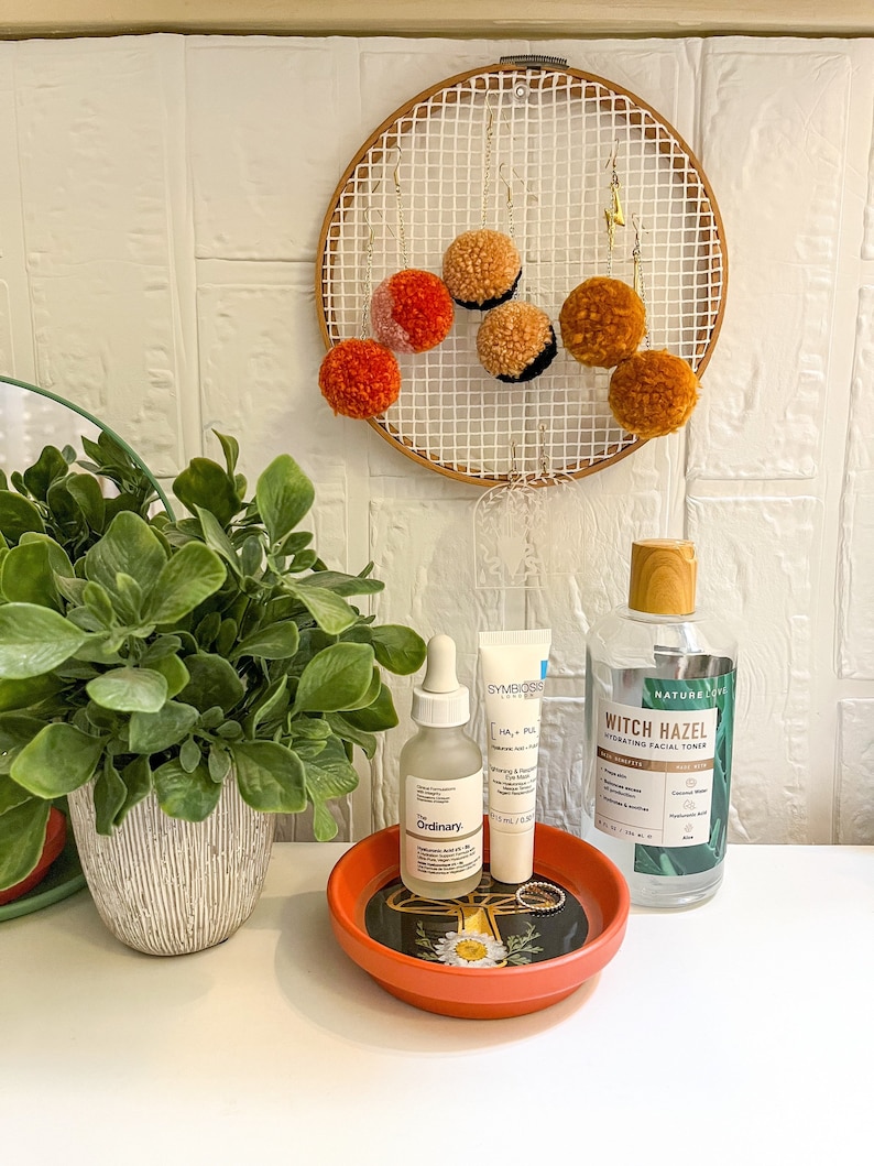May include: A small orange ceramic dish with a floral design holds three skincare products: a clear glass bottle with a dropper labelled "The Ordinary", a white tube labelled "Symbiosis HA + PL", and a clear glass bottle with a brown cap labelled "Nature's Love Witch Hazel Hydrating Facial Toner".
