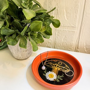 May include: A red ceramic dish with a black mushroom and white flower design. The dish is filled with a gold chain necklace and two gold rings.