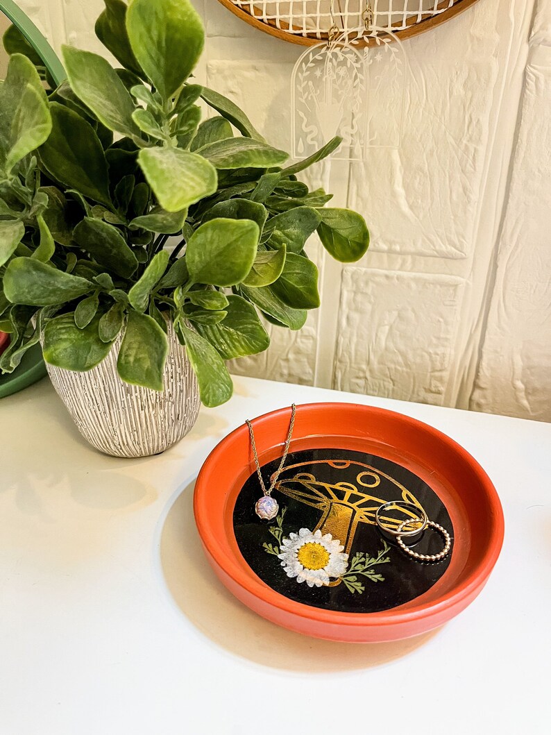 May include: A red ceramic dish with a black mushroom and white flower design. The dish is filled with a gold chain necklace and two gold rings.