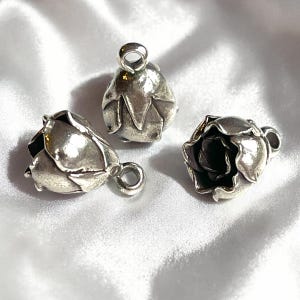 May include: Three silver-toned rose-shaped pendants with a loop for hanging. The pendants are detailed with petals and are displayed on a white, silky background. The pendants have a vintage or antique appearance.