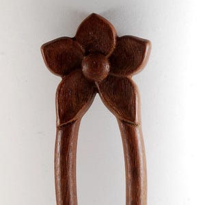 May include: A wooden hair fork with a flower design. The hair accessory is made of dark brown wood and features a carved flower at the top with two long, curved prongs. The hair fork is a decorative hair accessory.