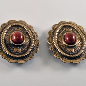Puede incluir: Un par de pendientes ovalados de estilo vintage. Cada pendiente presenta una piedra roja redonda central engastada en un marco de metal dorado detallado. El marco tiene un borde festoneado y patrones en relieve, creando una estética Western clásica.