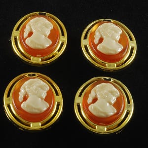 May include: Four gold-toned cameo earrings with a classic profile design. Each earring features a cream-colored cameo set against an orange background, framed by a gold border. The earrings are round and have a vintage aesthetic.