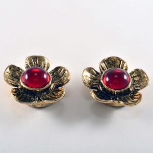 May include: A pair of flower-shaped earrings. Each earring features a gold-toned, petal-like design with a vibrant red, round gemstone at the center. The earrings are set against a white background.