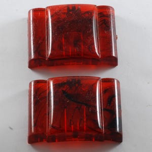 May include: Two rectangular, red-orange vintage hair clips with a marbled black pattern. Each clip has a central, raised rectangular section flanked by two smaller, vertical rectangular sections. The clips are made of a translucent material.