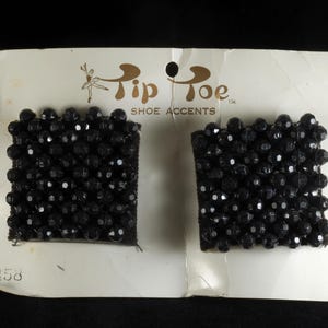 May include: Pair of black beaded shoe accents on a white card. The card reads "Tip Toe Shoe Accents" with a ballerina graphic. The accents are square-shaped and densely covered in small, faceted black beads. The card has a hole for hanging.