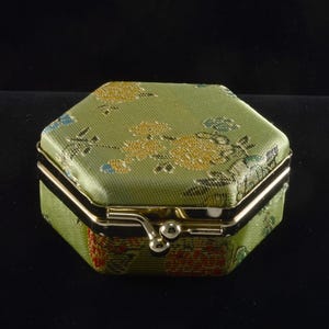 May include: A small, hexagonal jewelry box with a green floral pattern. The box has a gold-colored metal trim and clasp. The background is black.