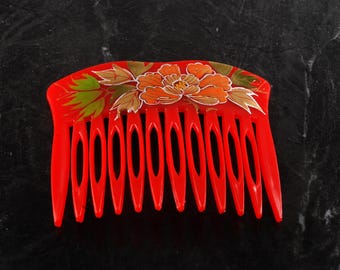May include: A vibrant red hair comb with a floral design. The comb features a large orange flower with gold and green accents. The teeth of the comb are long and evenly spaced.