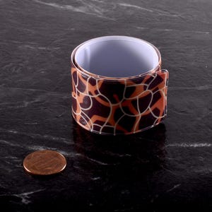 May include: A cylindrical slap bracelet featuring a dark brown and orange giraffe-like pattern with white outlines. The interior of the bracelet is white. A copper-colored United States penny is positioned next to the bracelet for scale.