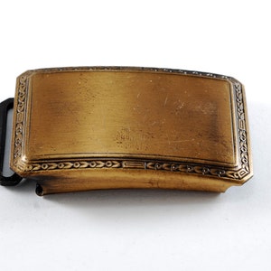 May include: A gold-toned metal belt buckle with a decorative border. The buckle is rectangular with rounded corners and has a smooth, polished surface.