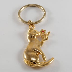 May include: Gold-colored keychain featuring a cat figurine holding a red flower. The cat is sitting with its tail curled around its body.