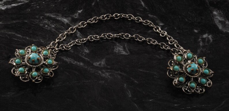 May include: A silver chain necklace with two turquoise flower-shaped pendants. Each pendant has a central turquoise stone surrounded by smaller turquoise stones.